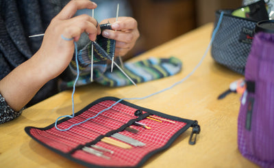 lifestyle visible^^A person knitting with their Pen Tool Wrap filled with knitting tools on the table next to them.