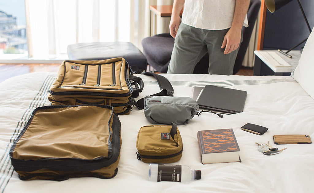 A person packing a Tri-Star with travel items, including a large Packing Cube, Spiff Kit, Stuff Sacks, a Book, a water bottle, a tablet, phone, and a wallet.