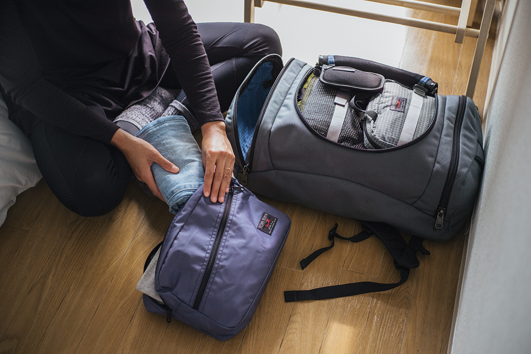 Person packs jeans into a Dusk Cerylon Packing Cube Shoulder Bag. A fully packed Aeronaut 45 in Steel Ballistic/Fjord 200 Halcyon sits to their left.