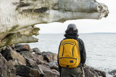 visible lifestyle^^Person with a yellow backpack standing on rocks by a body of water.