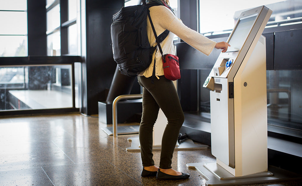 lifestyle visible^^Person wearing an Aeronaut 30 on their back as a backpack and an Everyday Cubelet across their body as they push a button on a check-in kiosk in an airport.