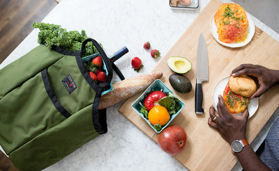 lifestyle visible^^A Moveable Feast on a counter holding WIne, rabe, a carton of strawberries, and a baguette next to a cutting board where a person is preparing some food.