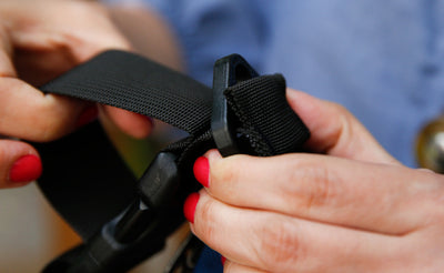 detail visible^^The edge of a stroller/wheelchair adapter strap through the gap between the webbing and the bottom of the d-ring shoulder-strap attachment point of a bag flared to keep it in place.