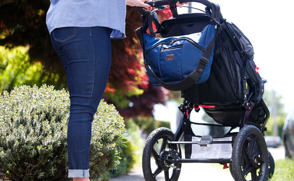 lifestyle visible^^A person pushing a stroller with a Deep Blue (dark blue) 525 Ballistic Parental Unit connected to the push handle of the stroller using the stroller/wheelchair adapter straps.