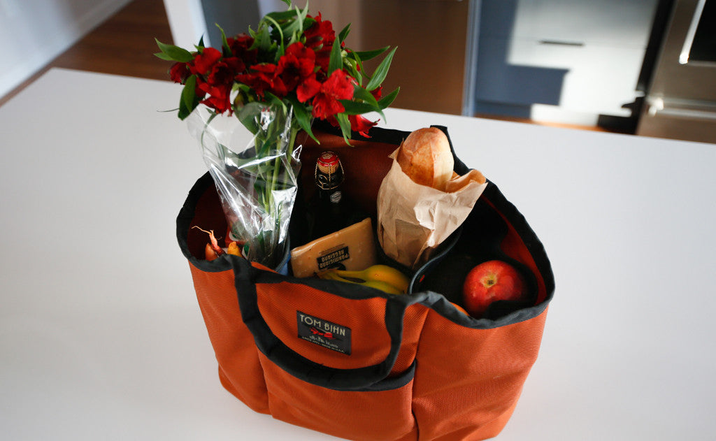 lifestyle visible^^A Moveable Feast holding flowers, vegetables, a beer, a baguette, some cheese, and an assortment of fruits.
