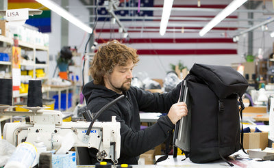 lifestyle visible^^Nik sitting at a sewing machine table in the TOM BIHN factory with the American and Rainbow flags on the wall in the background. He is pulling a 15" Dell laptop out of the laptop compartment of the Shadow Guide 33.