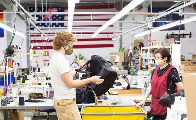 visible lifestyle^^Nik holding a prototype of the Shadow Guide 33 backpack in our Seattle factory. He is talking with Fong and both are wearing masks. Rainbow, U.S.A. and Earth flags are in the background.