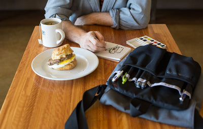 visible lifestyle^^Person creating a watercolor illustration in a cafe. Their Medium Cafe Bag and its Freudian Slip is on the table, along with a breakfast sandwich and cup of tea.