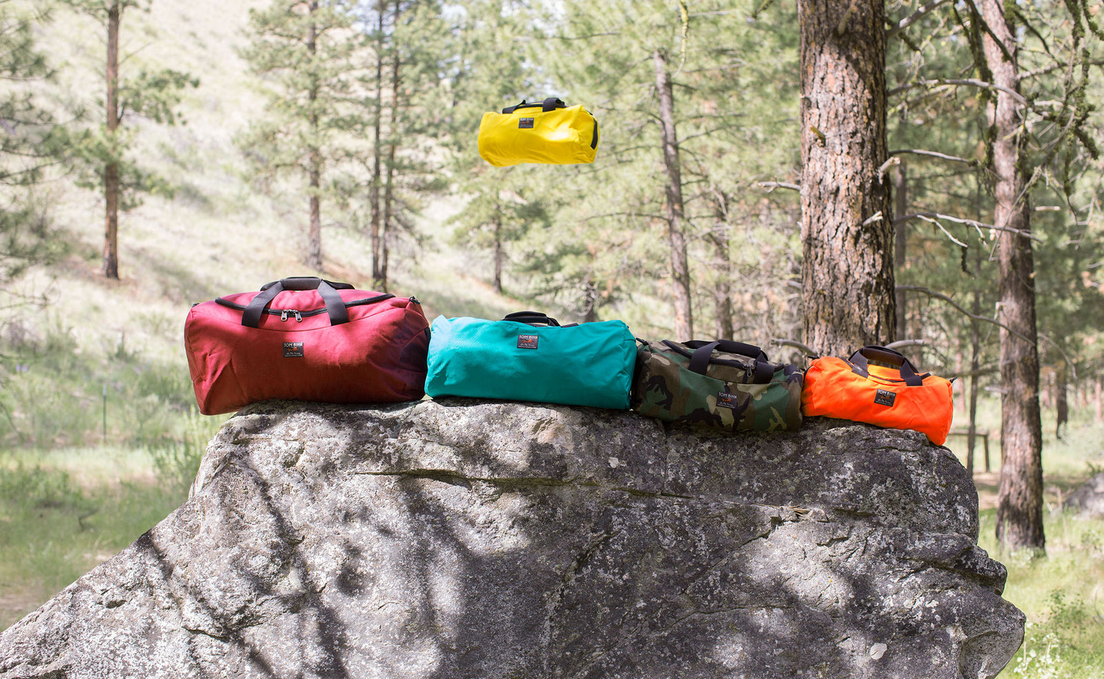 lifestyle  visible^^Various sizes and colors of Yeoman stacked on top of a large rock at a campsite in the woods. A Mini Yeoman in Canary is in the air behind the rock.