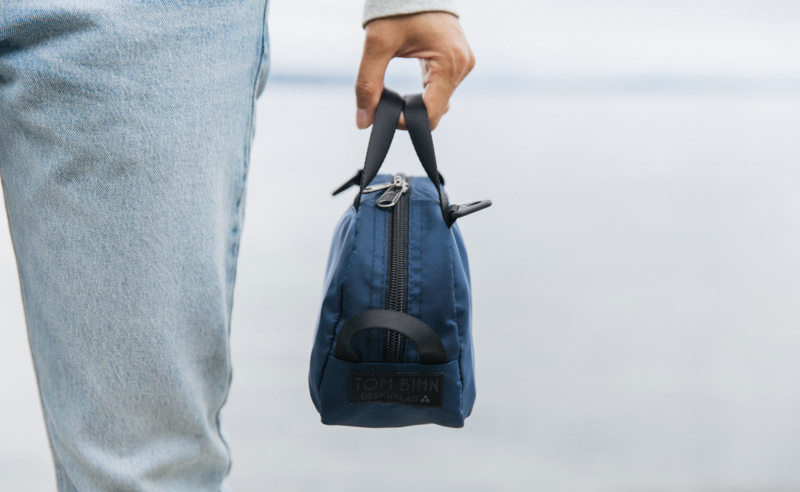 lifestyle visible^^ A person holding a Navy Parapack Grab Bag by it's grab handle showing the TOM BIHN Design Lab label. 