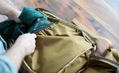 detail visible^^A person sliding a rain shell into the elasticized internal pocket of the main pocket of the Guide's Edition Synik 22.