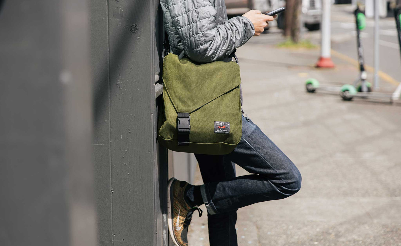 lifestyle visible^^Person wearing the Large Cafe Bag in 1000d Cordura Olive cross-body. Leaning against a wall, looking at their phone.