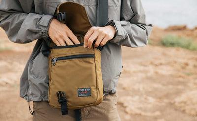 Person wearing the Small Cafe Bag in Coyote 210d ballistic. They are reaching in the main compartment.