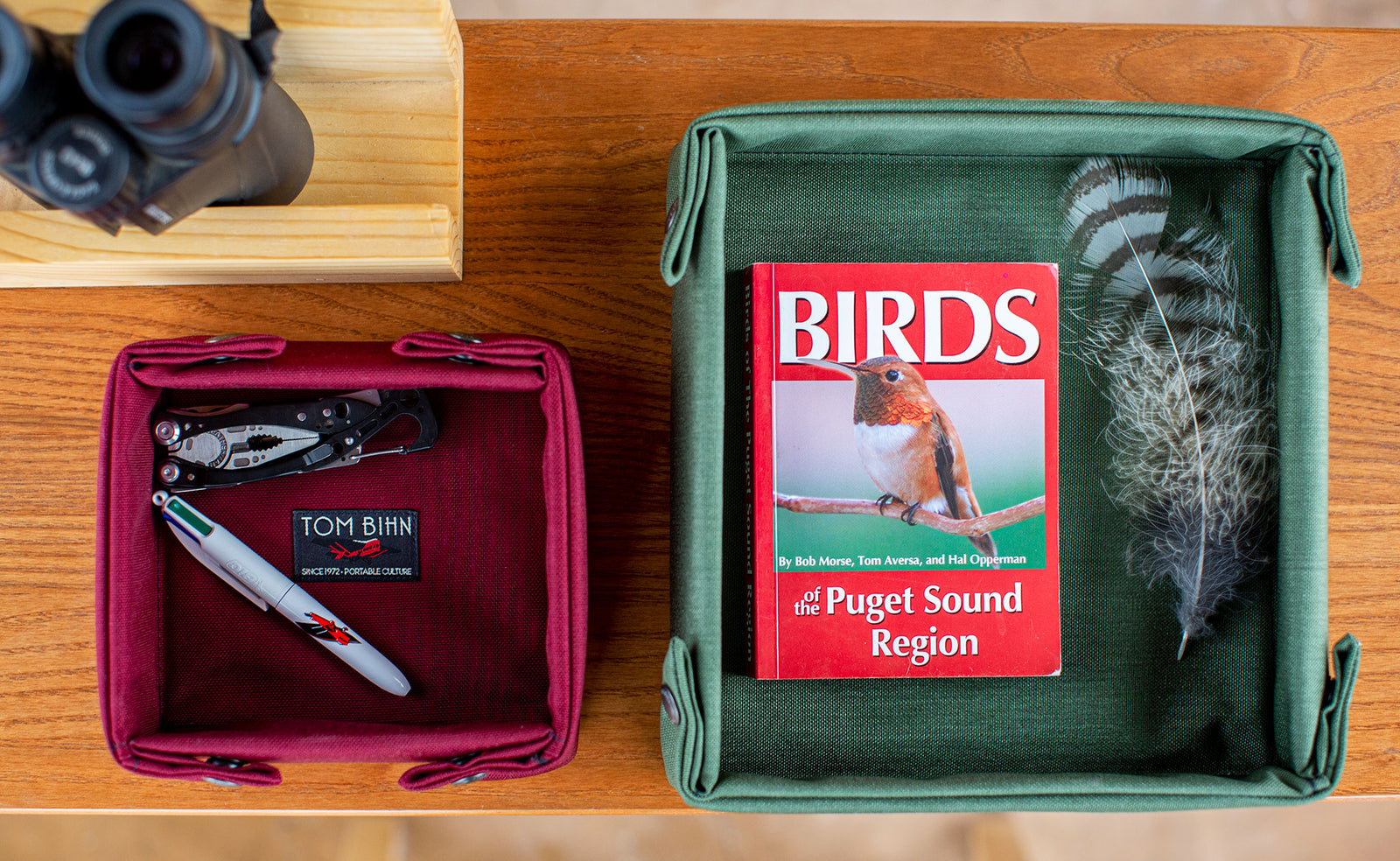 lifestyle visible^^Small Tray Bien in Red Blend with a Skeletool and a TOM BIHN writes-in-four-colors pen; Tray Bien, Medium, in Boreal with the "Birds of the Puget Sound Region" book and a bird's feather.