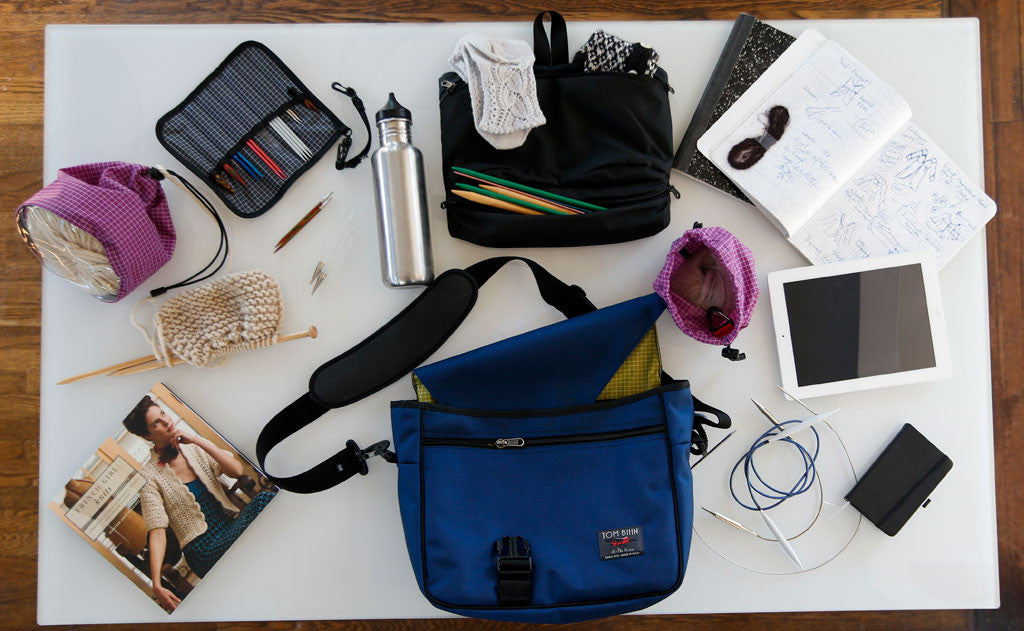 lifestyle visible^^The knitting loadout of a knitting including a Small and Medium Ultraviolet (purple with white grid) 200 Halcyon Yarn Stuff Sack, Maker's Freudian Slip in Black 210 Ballistic, and a Maker's Bag in Navy (blue) 1050 Ballistic with a Wasabi (green with white grid) 200 Halcyon interior, tablet, cable knitting needles, an in-progress project, pens, a water bottle, notebooks, and a knitting book.