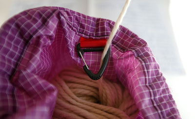 ^^A string of a skein of yarn being held in an Ultraviolet (purple with white grid) 200 Halcyon Yarn Stuff Sack pulled out of the sack using the interior clip as a guide for the yarn.