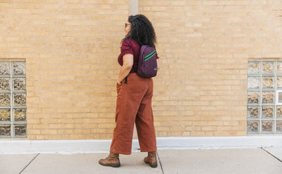 visible detail^^Person wearing the Zeitgeist backpack in Aubergine. They're standing outdoors in front of a brick building.